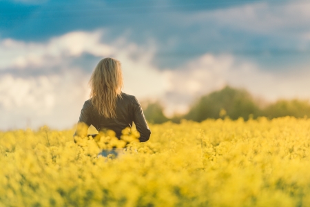 A lady with blonde hair and a black leather jacket is stood in a field of rapeseed flowers with her back to us looking towards a cloudy blue sky in the background.