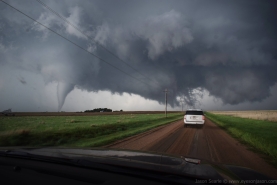 A photograph taken from a car windscreen of a dirt road with a white SUV driving towards a thin and ropey tornado, with a cone-shaped tornado on the left of the screen. The image looks dramatic.