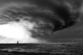 A black and white image of a mesocyclone, which looks like it is spiraling to the ground. The horizon is dotted with wind-turbines.